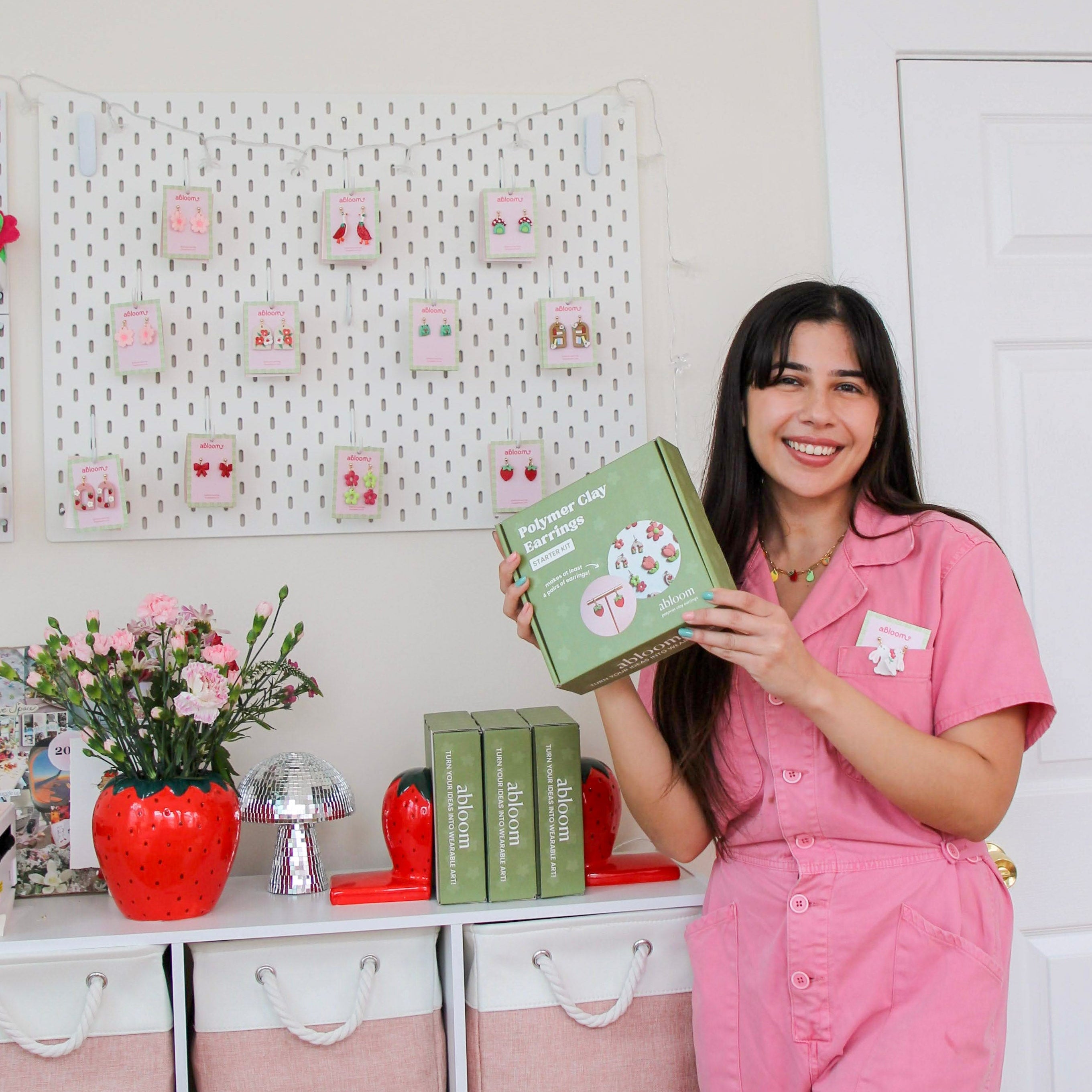 Woman in pink outfit holding a diy kit box in a room with earrings on the wall and shelves.