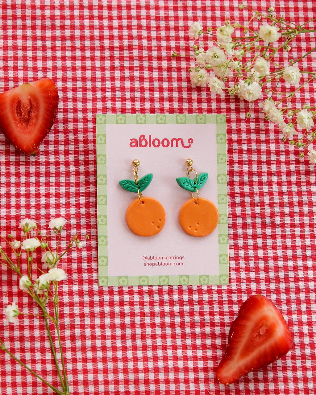 Pair of orange earrings with green leaves on a 'abloom' branded card, placed on a red and white checkered background with strawberries and flowers.