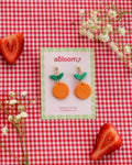 Pair of orange earrings with green leaves on a 'abloom' branded card, placed on a red and white checkered background with strawberries and flowers.