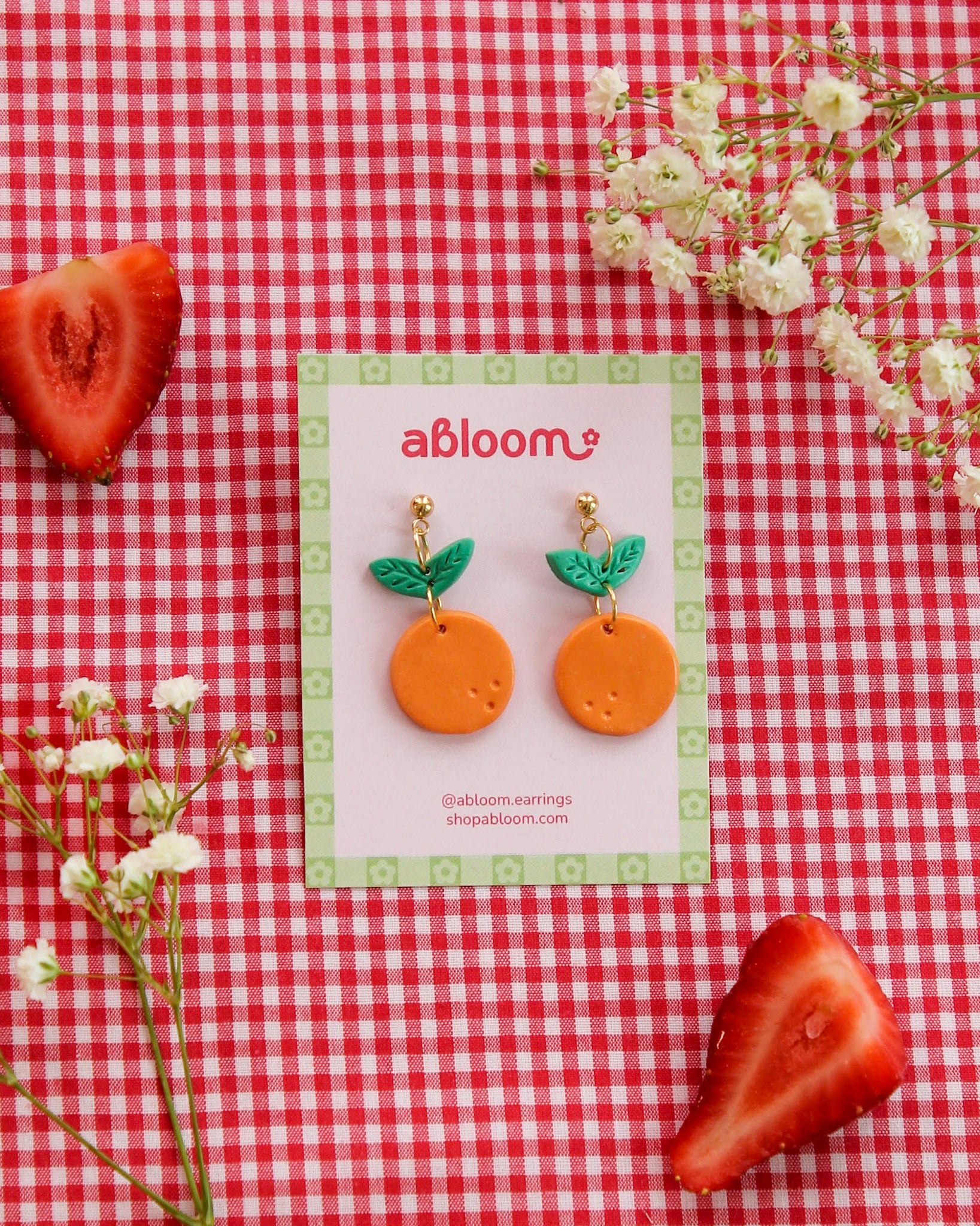 Pair of orange earrings with green leaves on a 'abloom' branded card, placed on a red and white checkered background with strawberries and flowers.