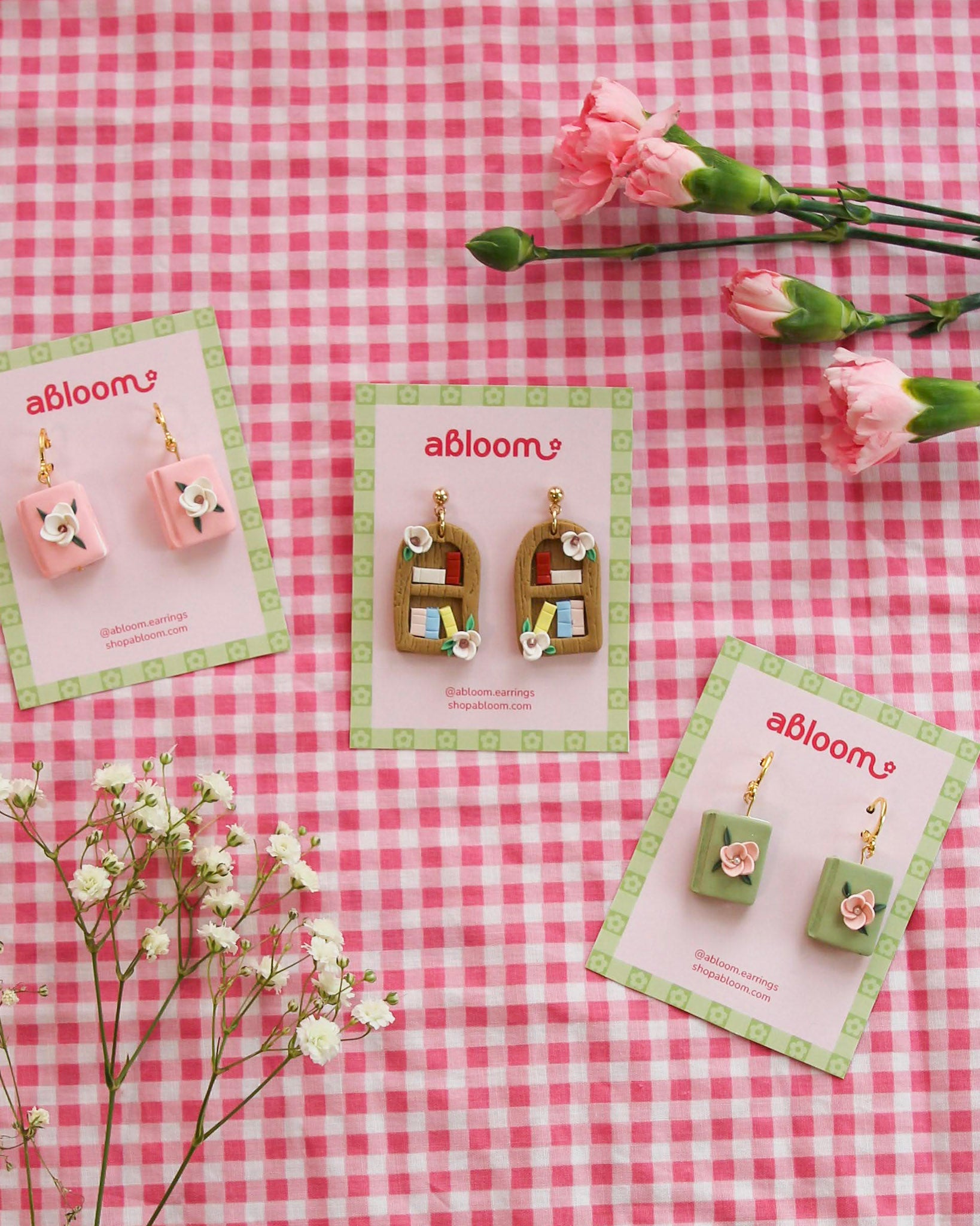 Three pairs of earrings on cards with 'abloom' branding on a pink checkered background with flowers.