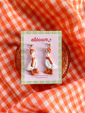 A pair of white goose-shaped earrings wearing a matching orange beanie, scarf, and boots.
