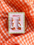 A pair of white goose-shaped earrings wearing a matching orange beanie, scarf, and boots.
