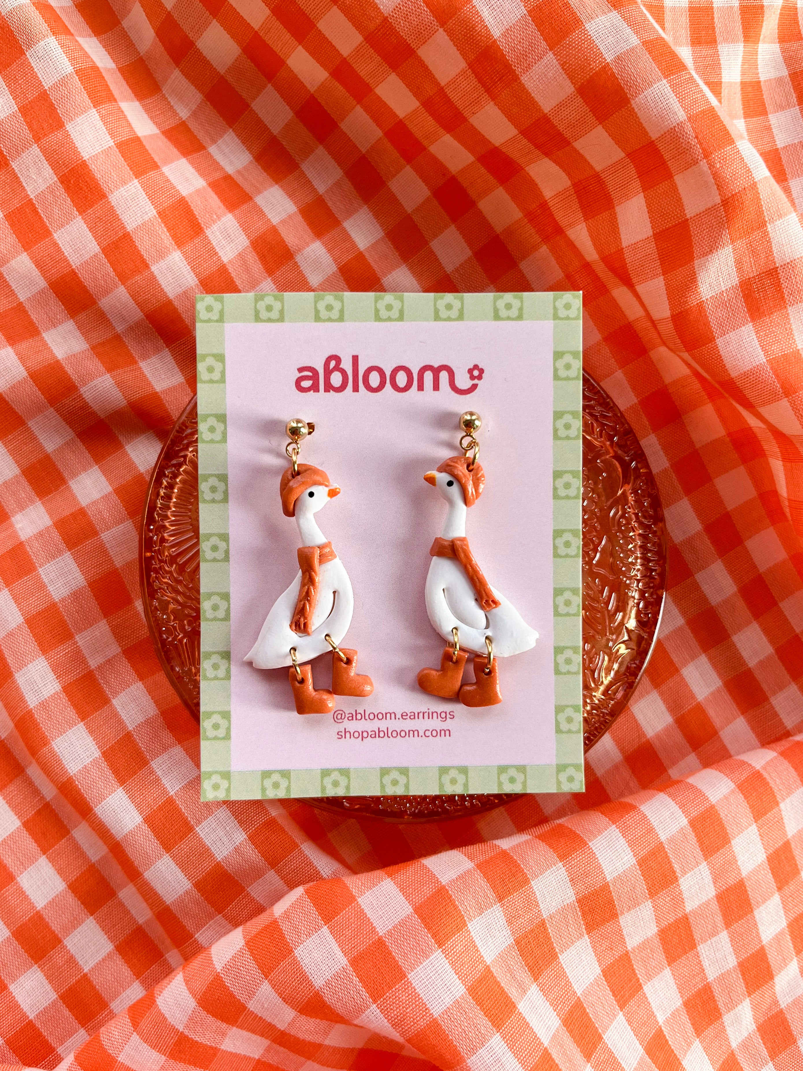 A pair of white goose-shaped earrings wearing a matching orange beanie, scarf, and boots.