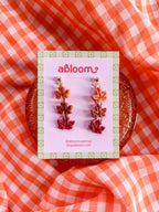 A pair of dangling maple leaf earrings displayed on a pink card.