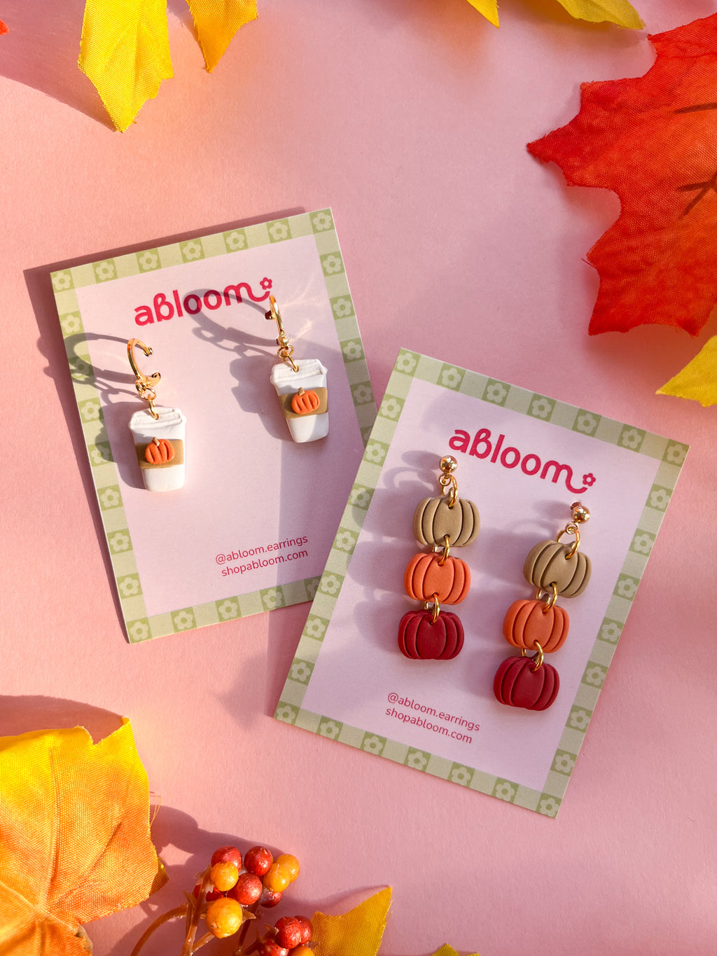 Two pairs of abloom earrings displayed on branded cards over a pink background with autumn leaves. One pair features a white pumpkin spice latte to go cup, and the other features stacked pumpkin charms in beige, orange, and red.