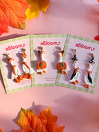 Three pairs of autumn-themed goose earrings. The earrings feature white, orange, black, and green details, styled with colorful fall leaves on a pink background.