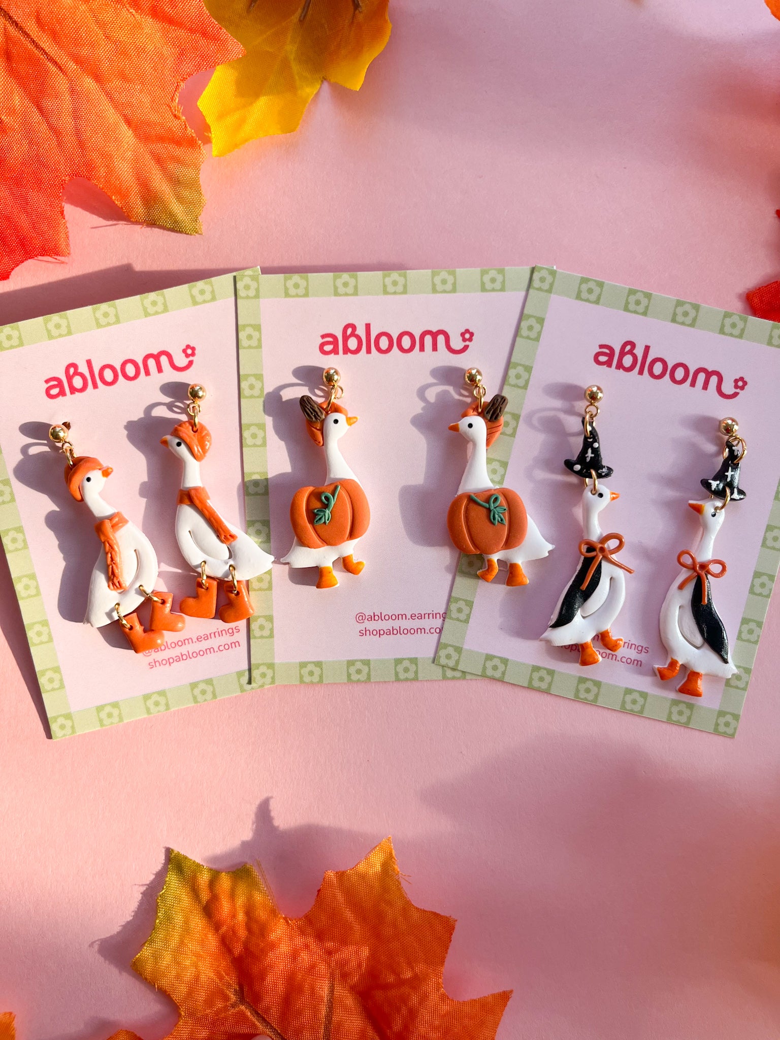 Three pairs of autumn-themed goose earrings. The earrings feature white, orange, black, and green details, styled with colorful fall leaves on a pink background.