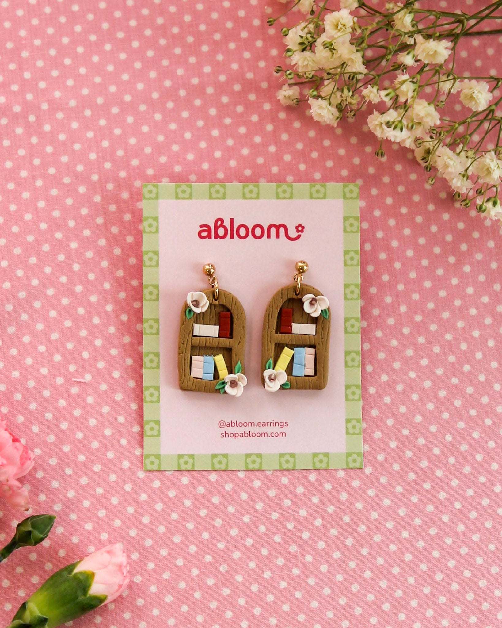 Bookshelf-themed earrings on a 'abloom' branded card with flowers on a pink polka dot background