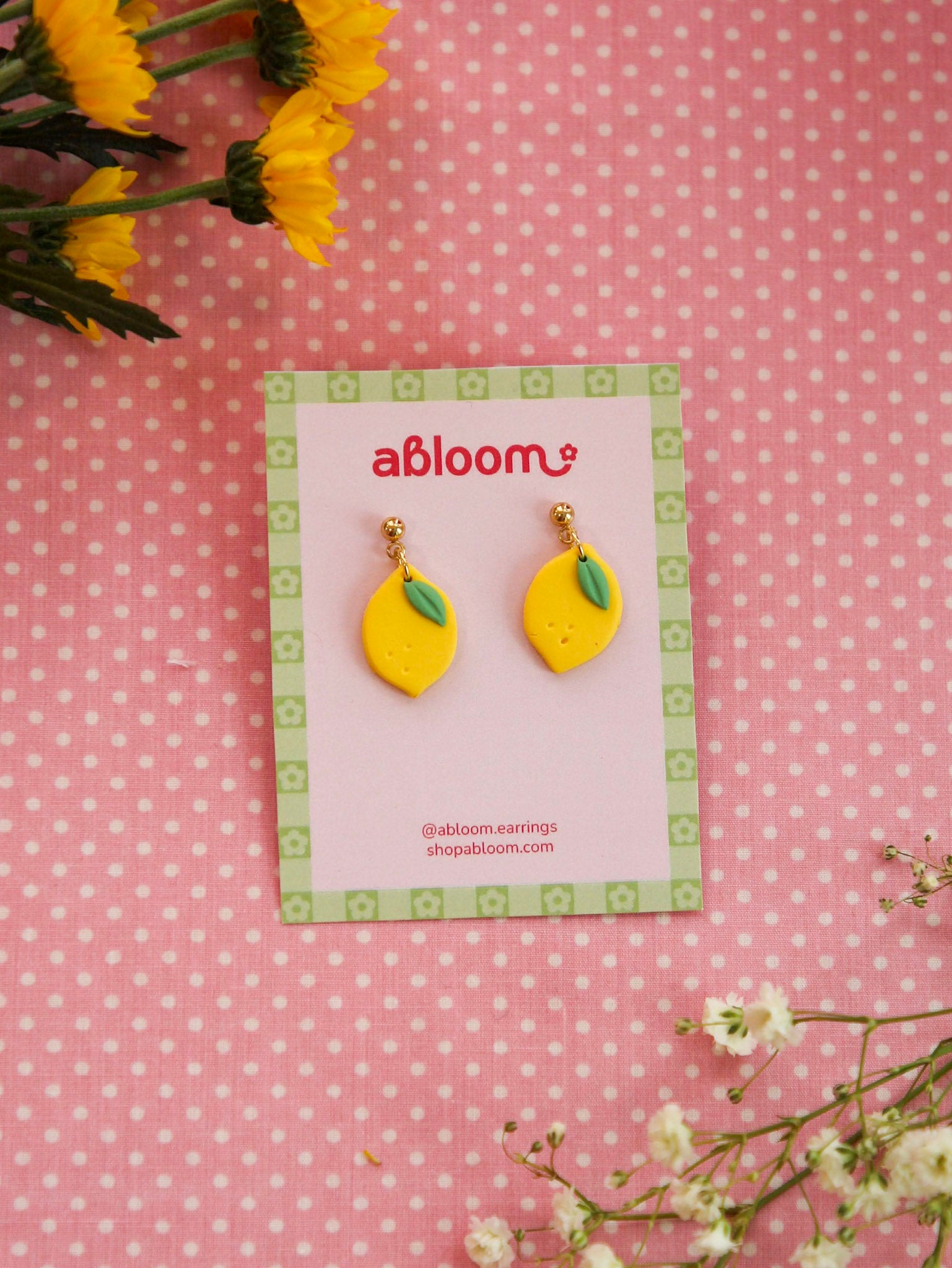 Yellow lemon-shaped earrings on a 'abloom' branded card with flowers around on a pink polka dot background
