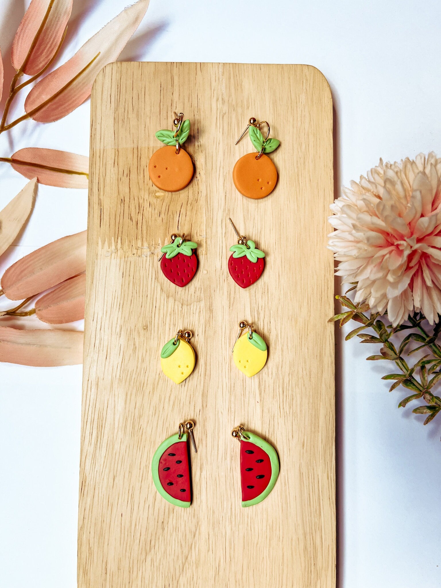 Fruit-themed earrings on a wooden board with flowers in the background