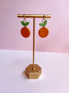 orange-shaped earrings with green leaves on a gold stand against a pink background