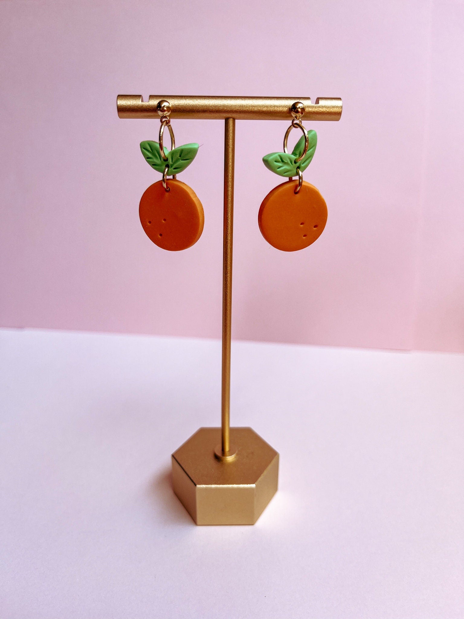 orange-shaped earrings with green leaves on a gold stand against a pink background
