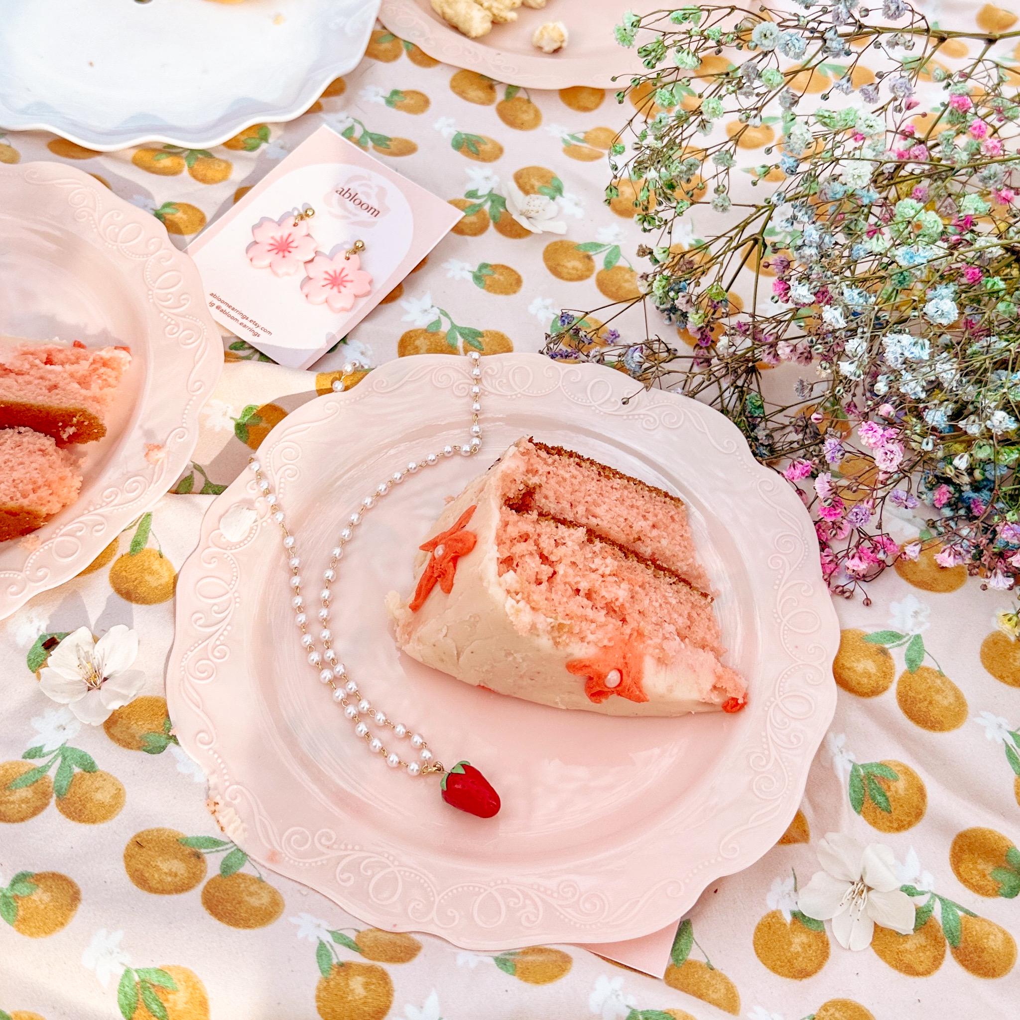 Slice of cake on a decorative plate with a strawberry pearl necklace and flowers on a patterned tablecloth