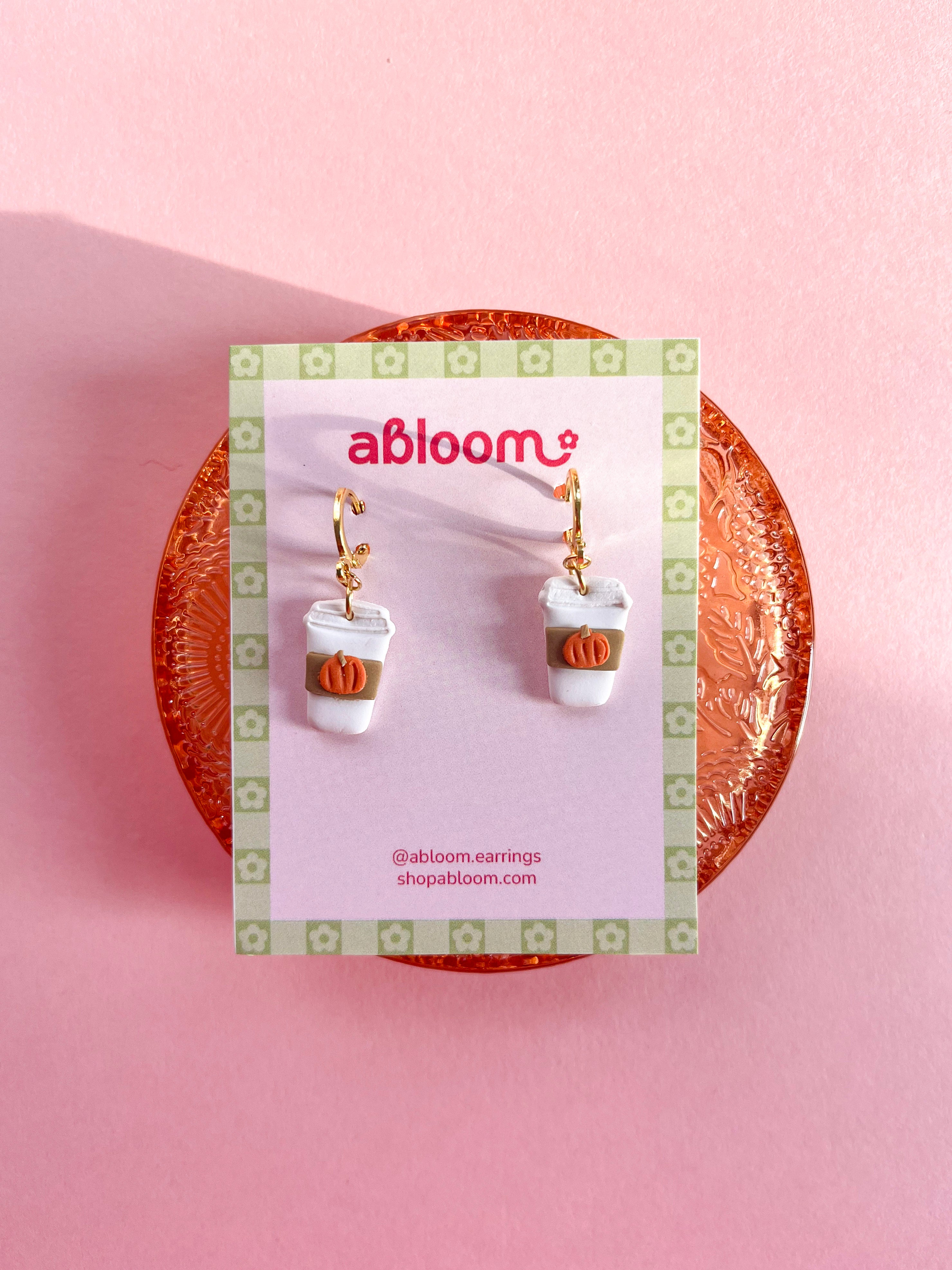 A pair of dangling earrings in the shape of a to go coffee cup, with a beige sleeve and tiny orange pumpkin. 