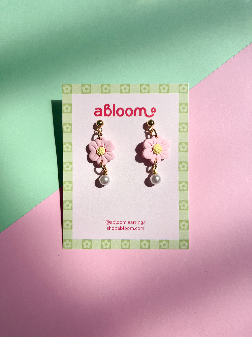 Pair of pink flower earrings with pearl centers on a 'abloom' branded card against a green and pink background.