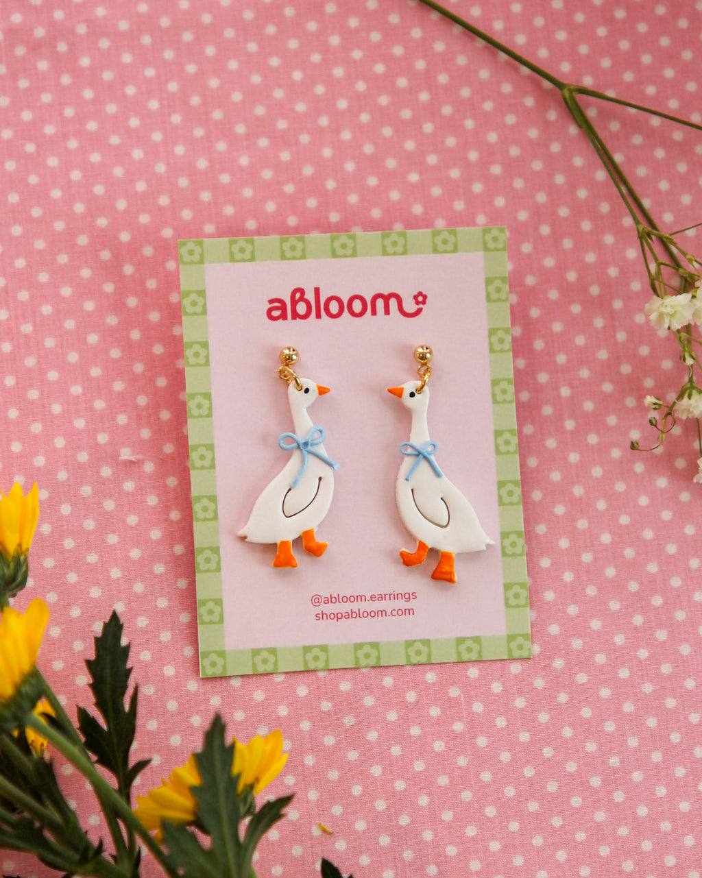 Pair of goose earrings on a 'abloom' branded card with flowers in the background.