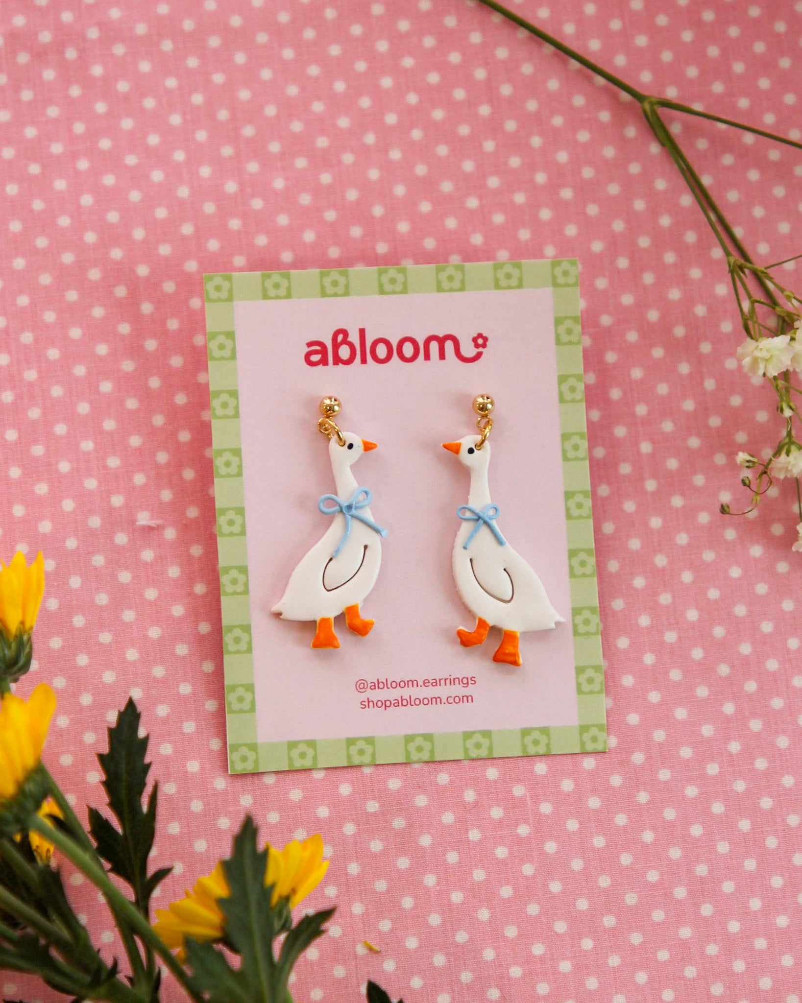Pair of goose earrings on a 'abloom' branded card with flowers in the background.