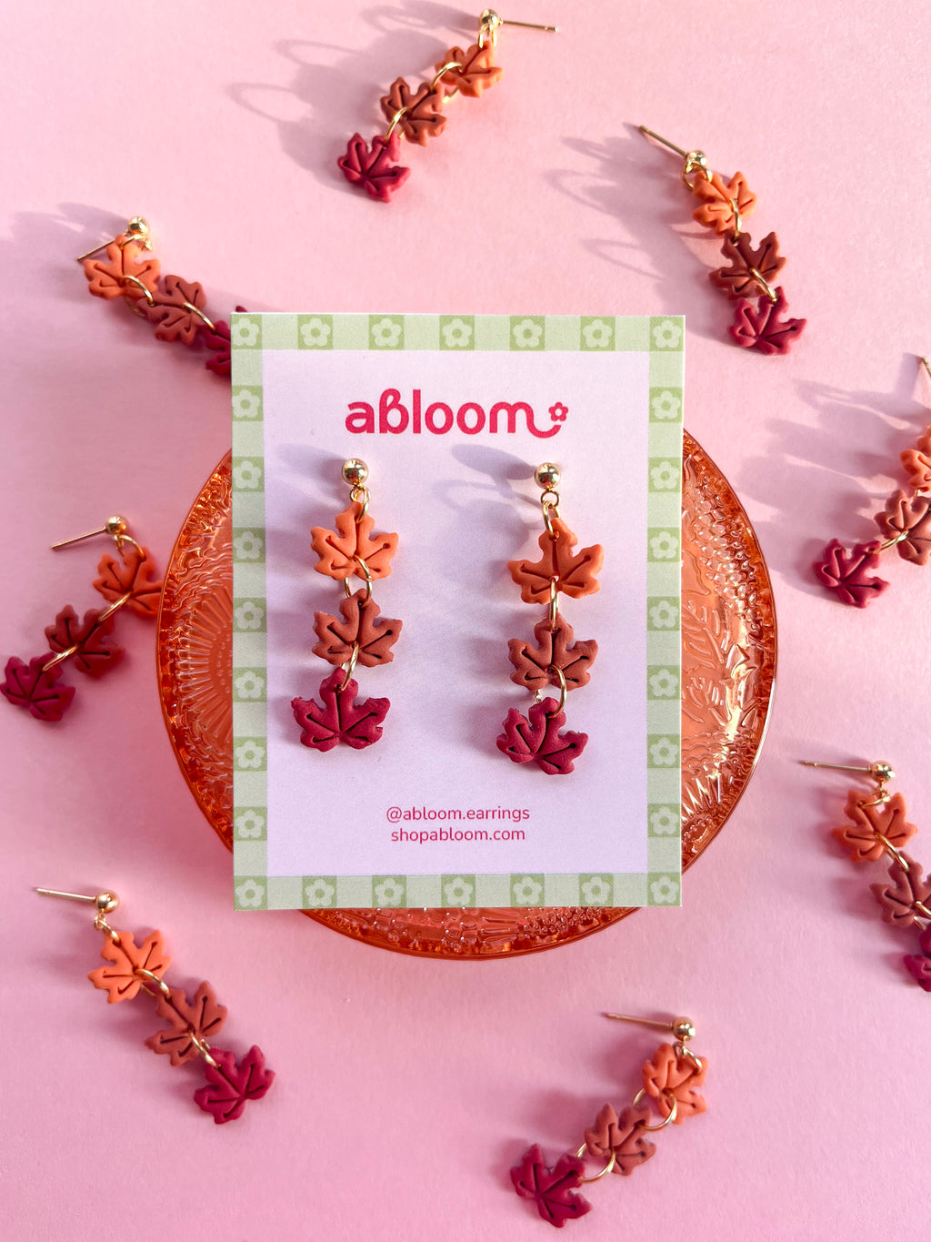 A pair of dangling leaf earrings displayed on a branded card, surrounded by additional matching earrings.