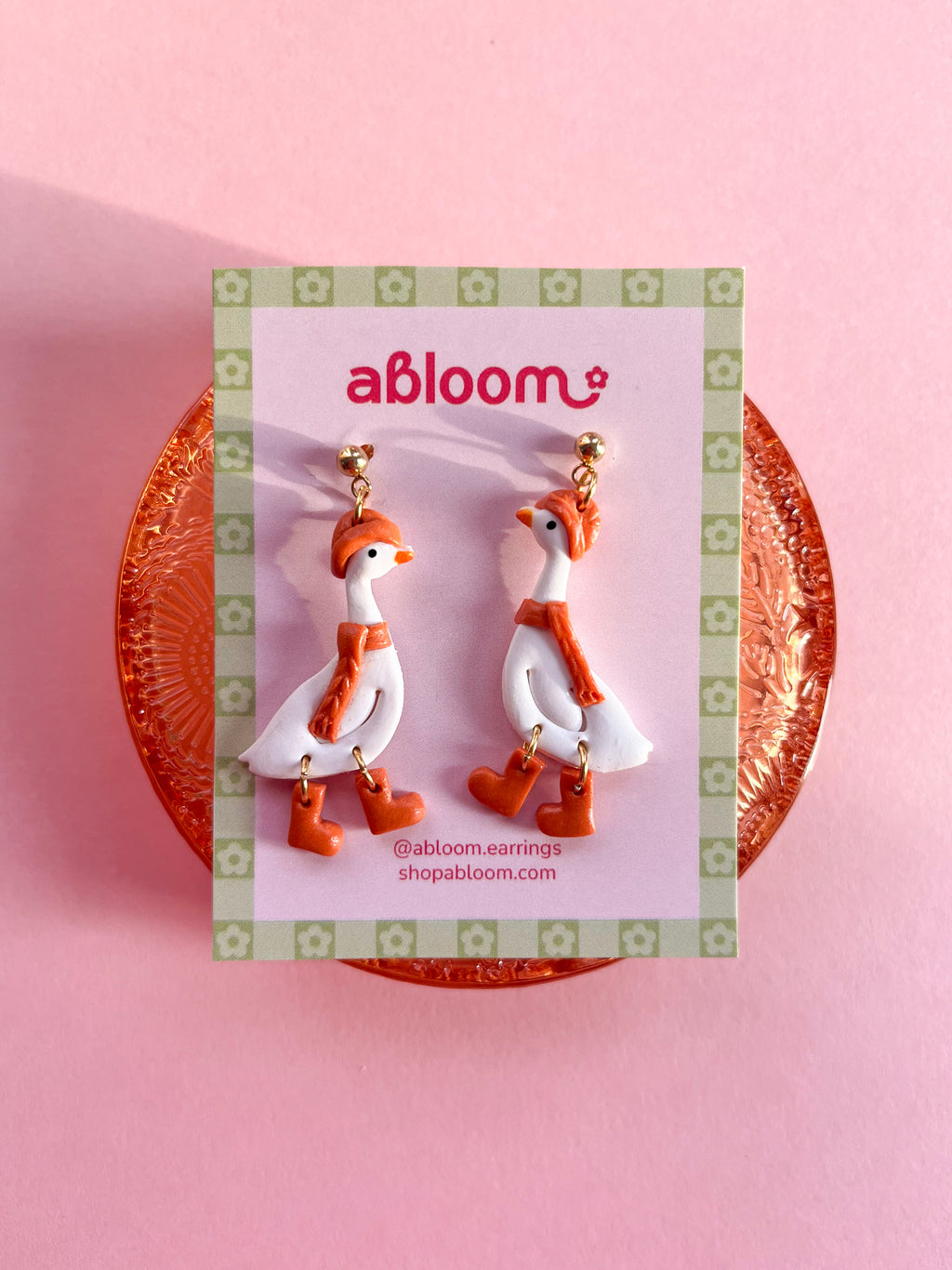 A pair of white goose-shaped earrings wearing a matching orange beanie, scarf, and boots.