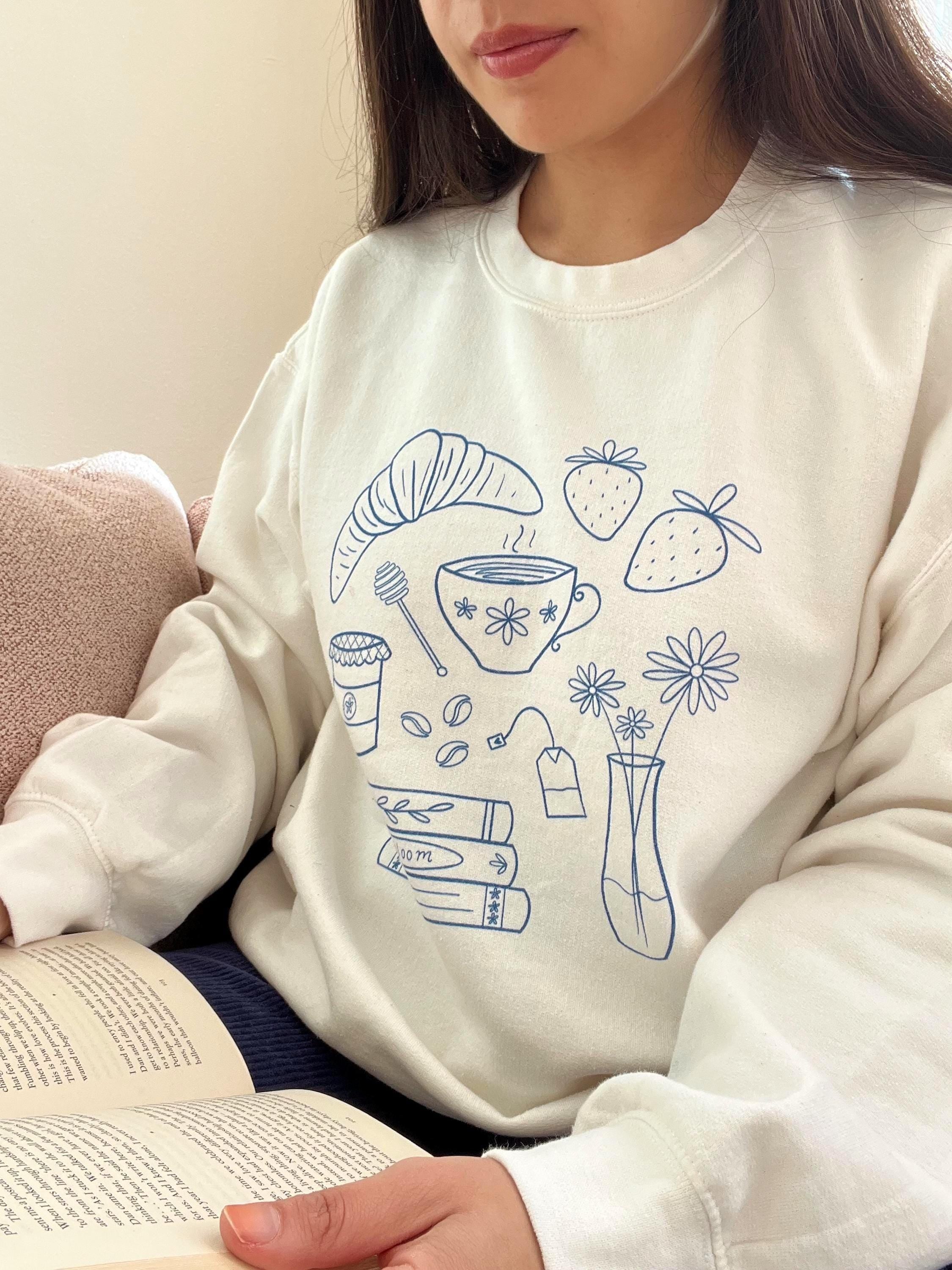 Person wearing a white sweatshirt with blue illustrations, holding an open book.