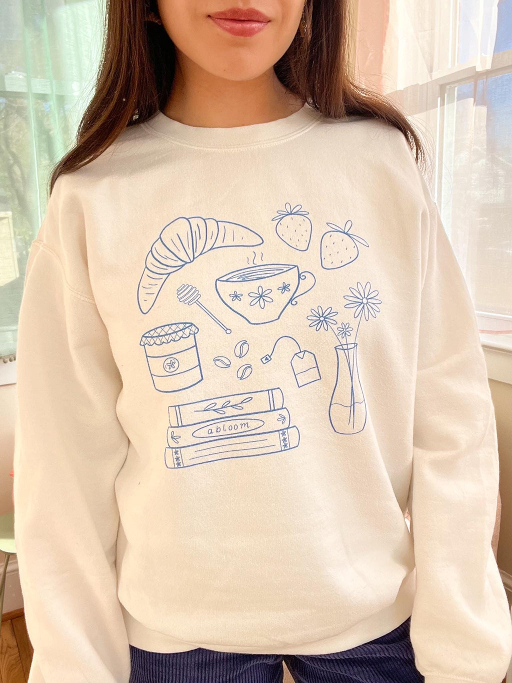 Person wearing a cream sweatshirt with blue illustrations of coffee, books, and other items.