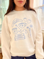 Person wearing a cream sweatshirt with blue illustrations of coffee, books, and other items.