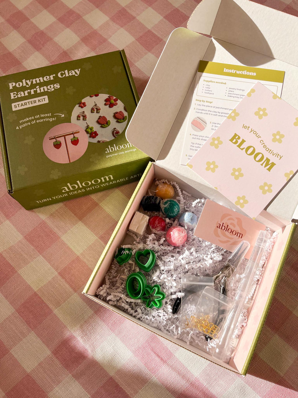 Open box containing polymer clay earrings kit with 'abloom' branding on a checkered tablecloth.