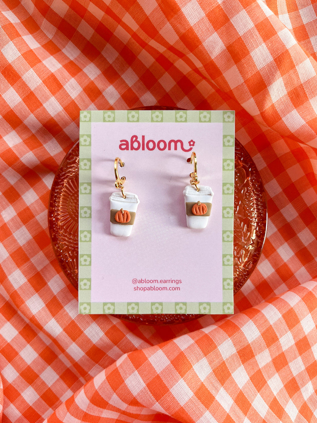 A pair of dangling earrings in the shape of a to go coffee cup, with a beige sleeve and tiny orange pumpkin. 