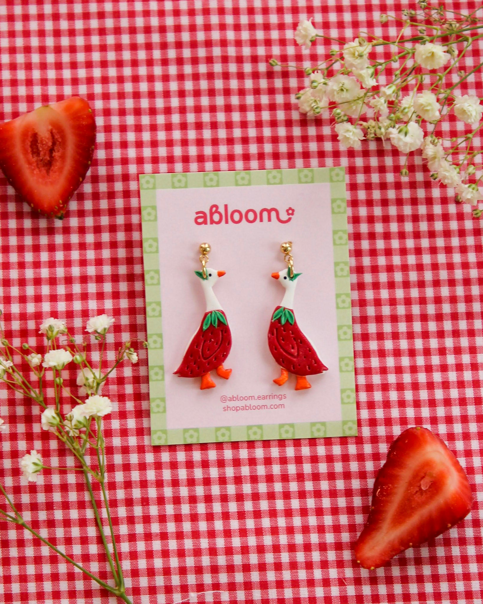 Strawberry goose earrings on a 'abloom' branded card with strawberries and flowers on a red checkered background