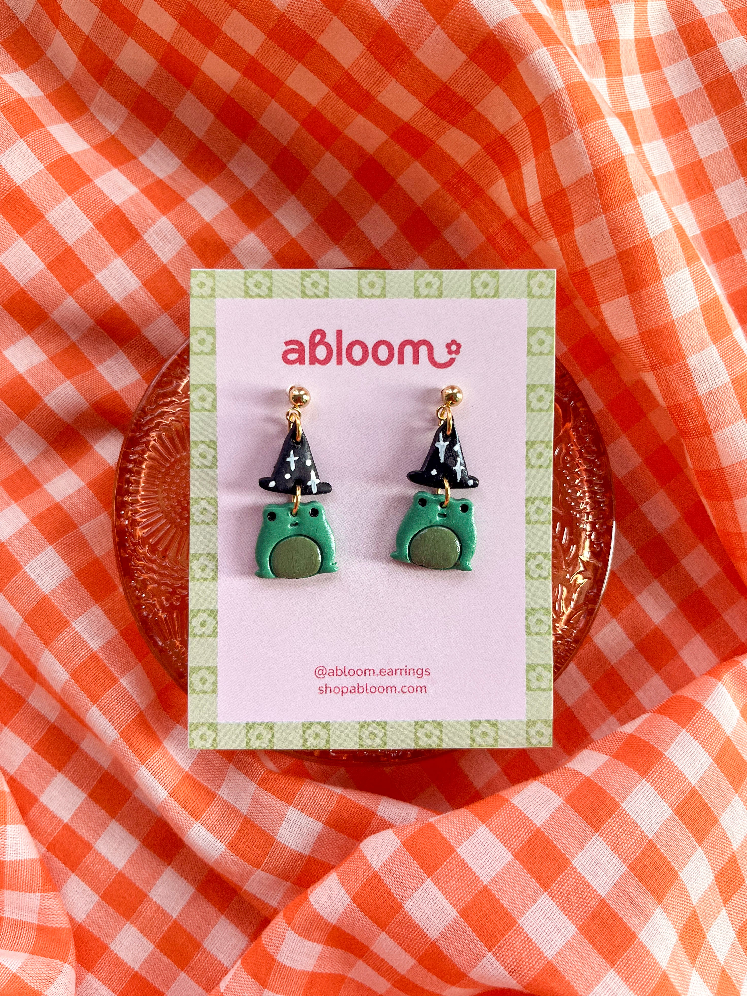 A pair of small frog-shaped earrings wearing a witch hat